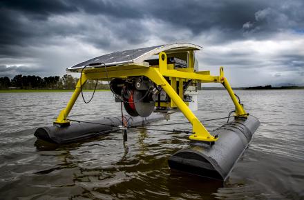 A structure with solar panels on top and four yellow legs sits on two floating rectangular bouys. The robot is on a lake, with dark clouds in the background