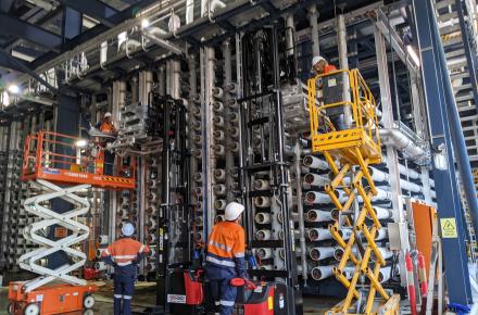 Two men on cherry pickers, supported by two workers on the ground, replace reverse osmosis membranes at the desalination plant