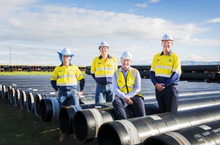 Four men in high vis shirts with hardhats stand between a row of black pipelines