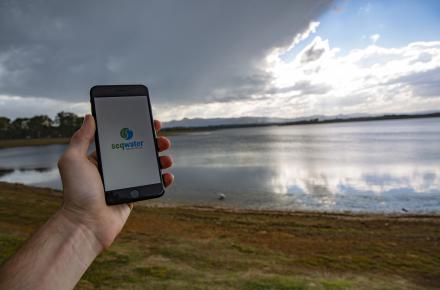 A hand hold a mobile phone screen with the Seqwater logo on it, in front of an approaching storm