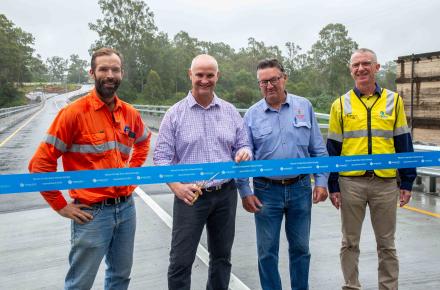 Four men stand in a line in front of a blue ribbon with a bridge behind them.