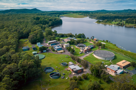 An aerial photo overlooking a small water treatment plant comprised of small orange brick buildings and a large round concrete tank, on the shores of a blue grey lake