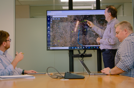 Three men in business shirts meeting around a large screen, with an aerial map on it