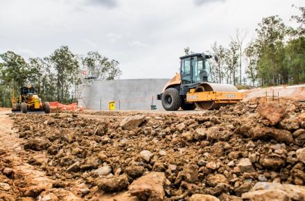 A construction site with heavy machinery, including a steamroller in the foreground and a yellow grader in the background